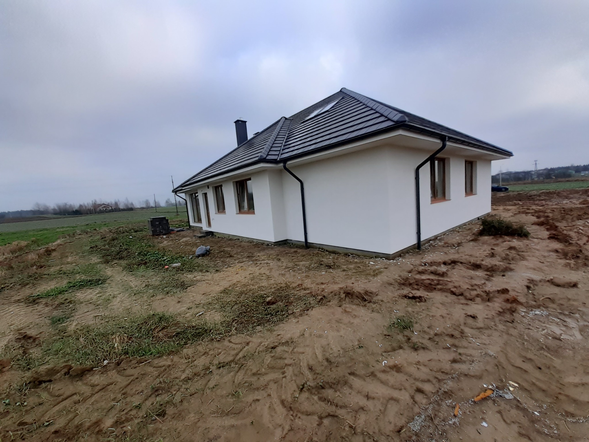 Nowoczesny dom jednorodzinny z białą elewacją i ciemną dachówką, widok z boku na tle pochmurnego nieba i przygotowanego terenu.