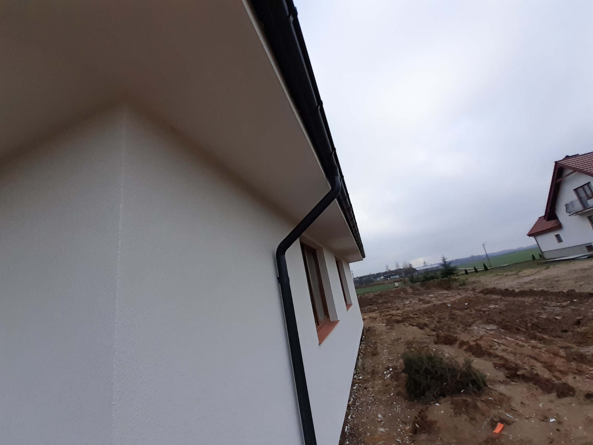 Fragment nowo wykończonej elewacji domu z widoczną strukturą tynku, czarną rynną i oknami z brązowymi ramami, na tle niezagospodarowanego terenu.