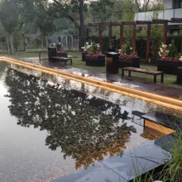 Podświetlony, prostokątny zbiornik wodny z kamiennym dnem, otoczony drewnianym tarasem z donicami kwiatowymi i ławkami, w tle drzewa i nowoczesny budynek.