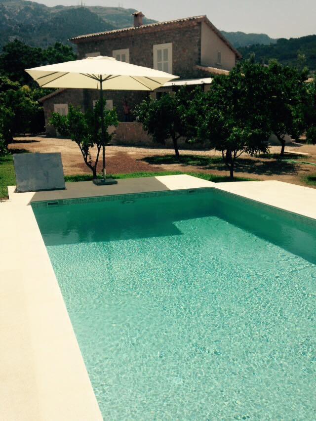 Turkusowa woda w prostokątnym basenie z jasnym obramowaniem, w tle dom z kamienia i góry, osłonięty białym parasolem.