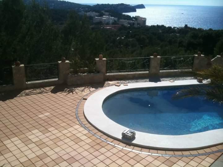 Owalny basen z błękitną wodą i jasnym obrzeżem, otoczony terakotową kostką, z widokiem na morze i zabudowania na wzgórzu.