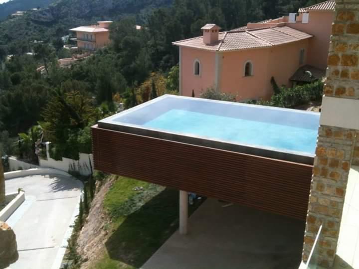 Nowoczesny basen bez krawędzi, z drewnianą obudową, wsparty na filarach, z widokiem na górzysty krajobraz i zabudowę mieszkalną w tle.