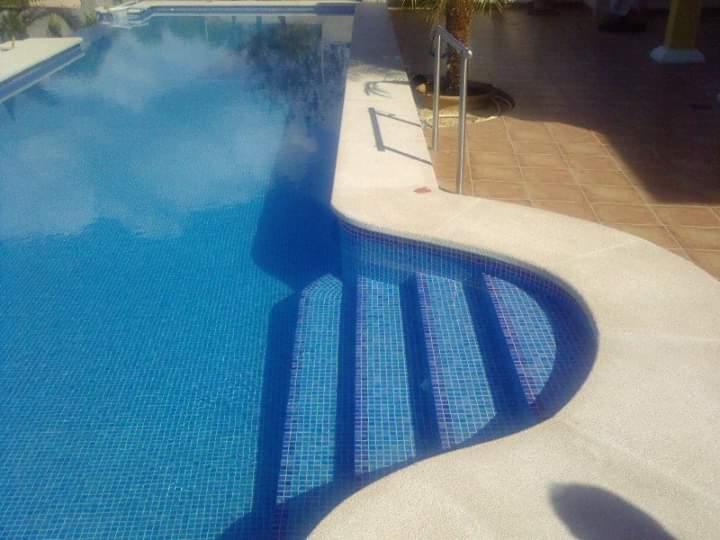 Błękitny basen z mozaiką i schodami, otoczony jasnym, teksturowanym obrzeżem, z widoczną metalową poręczą i fragmentem terakoty w tle.