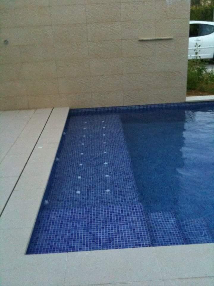 Niebieski basen wyłożony mozaiką z płytkimi schodami, obok ściana z beżowych płytek, w tle biały samochód.