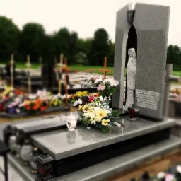 Szary nagrobek z rzeźbą kobiety w niszy, ozdobiony kwiatami i lampionami, na cmentarzu z innymi grobami w tle.