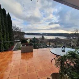 Błyszczący, pomarańczowy taras z widokiem na jezioro, czarne barierki, donica z kwiatami i stolik z lampionem.