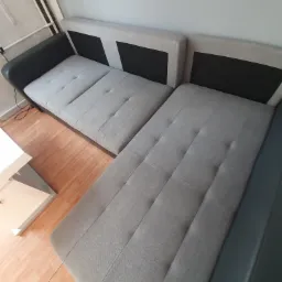 Szary narożnik z tapicerowanymi siedziskami i czarnymi elementami, stojący na drewnianej podłodze obok białego stolika kawowego.