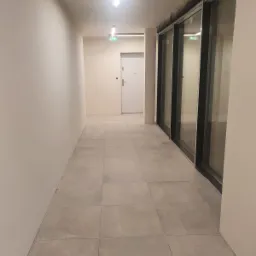 Wnętrze korytarza z jasnymi ścianami, szarą podłogą z płytek i rzędem okien z czarnymi ramami po prawej stronie, oświetlone górnym światłem.