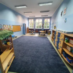 Przestronna sala przedszkolna z ciemnoniebieskim dywanem, drewnianymi regałami i szafkami, niebieskimi ścianami oraz oknami z widokiem na drzewa.