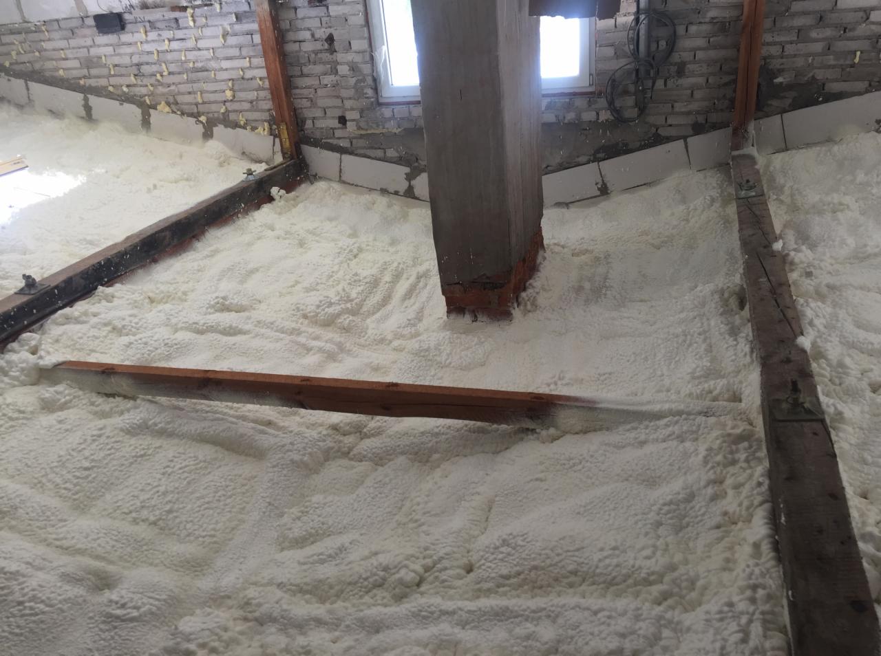 Poddasze z widoczną warstwą izolacyjną z pianki, drewniane belki konstrukcyjne częściowo zakryte pianą, fragment ściany z cegły i okno w tle.