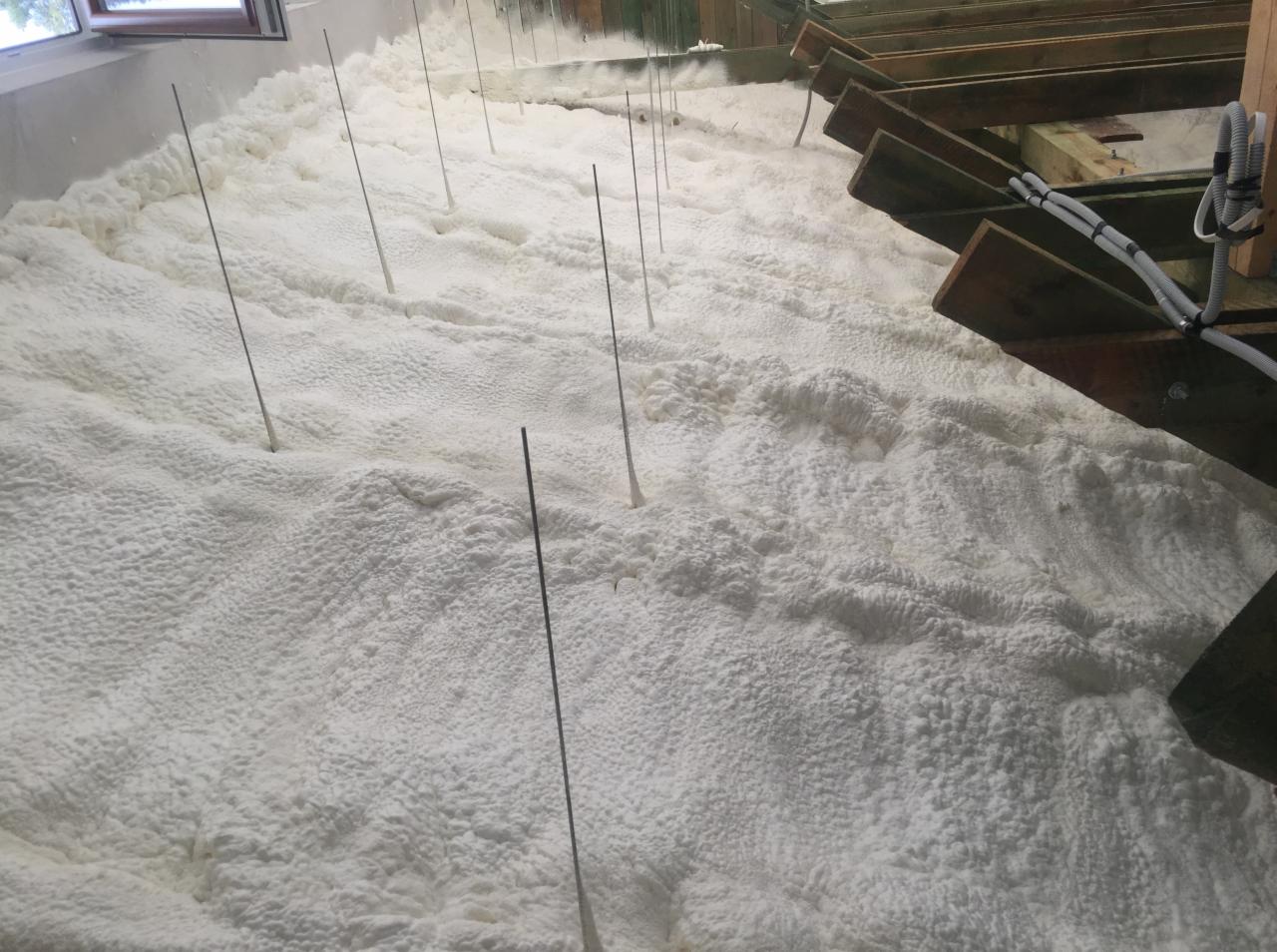 Świeżo nałożona, biała izolacja natryskowa na poddaszu, widoczne drewniane belki i wystające metalowe pręty stabilizujące.