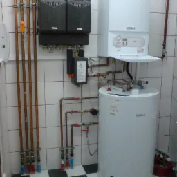 Biały, wiszący piec gazowy marki Vaillant, podłączony do zasobnika wody i rozdzielaczy z rurami miedzianymi, zainstalowany w pomieszczeniu z biało-czarną szachownicą na podłodze i białymi kafelkami...