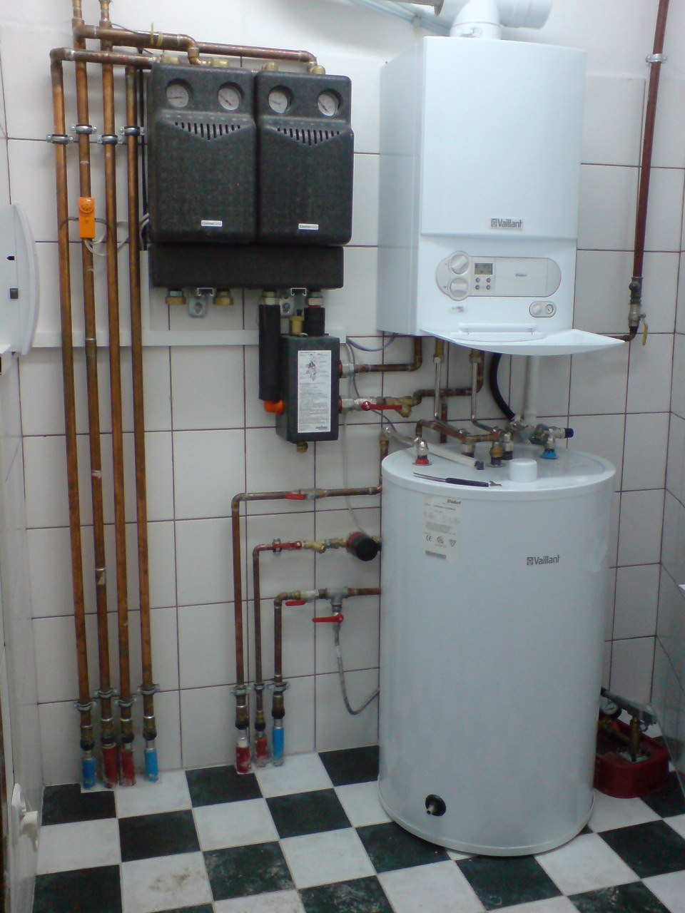 Biały, wiszący piec gazowy marki Vaillant, podłączony do zasobnika wody i rozdzielaczy z rurami miedzianymi, zainstalowany w pomieszczeniu z biało-czarną szachownicą na podłodze i białymi kafelkami...