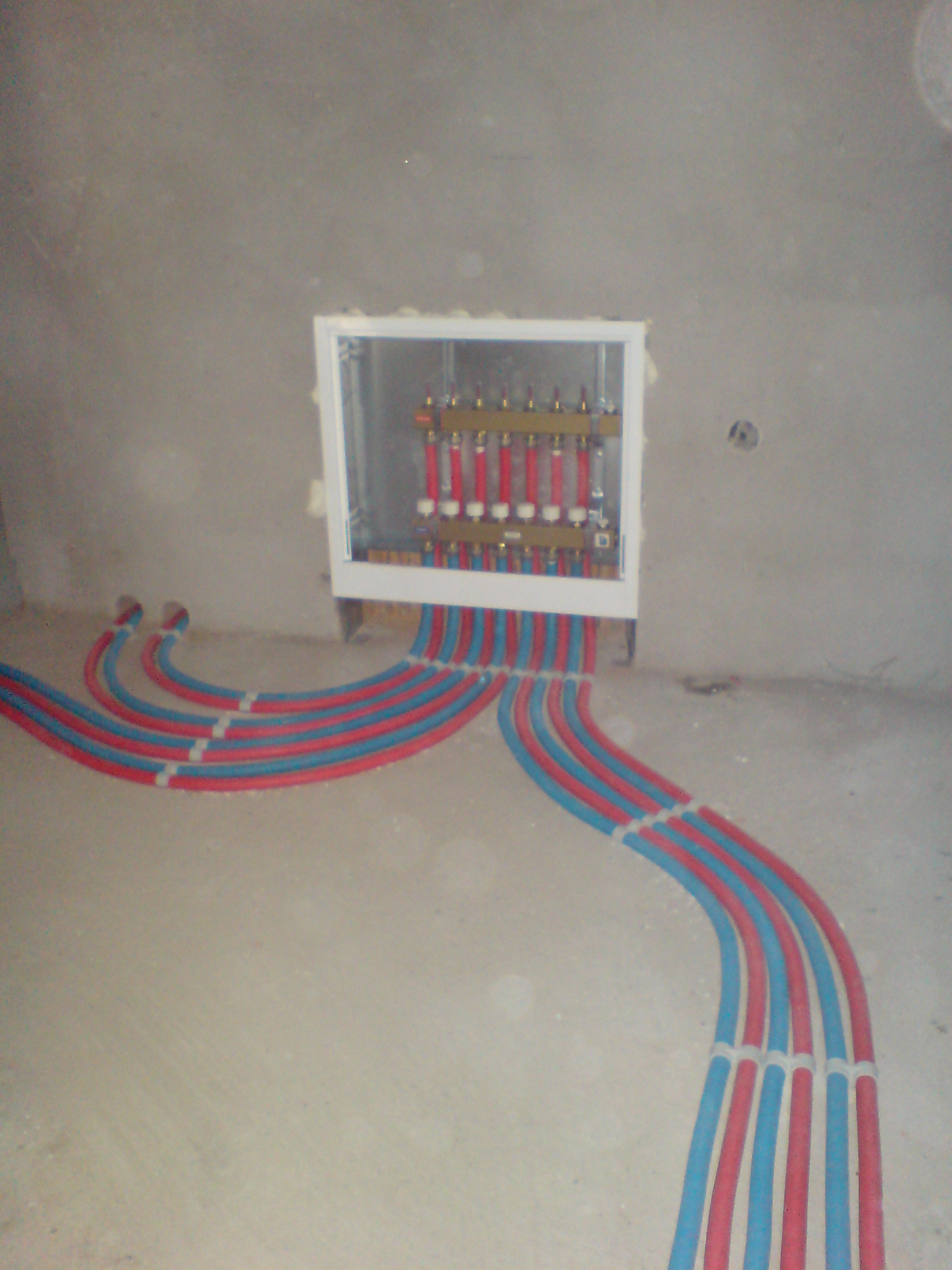 Rozdzielacz ogrzewania podłogowego z rurami czerwono-niebieskimi wychodzącymi ze ściany, surowy stan wykończenia.