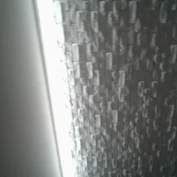 Ściana wykończona tapetą imitującą mur z cegieł w odcieniach szarości, oświetlona z boku światłem, z widocznym fragmentem gładkiej ściany po lewej stronie.