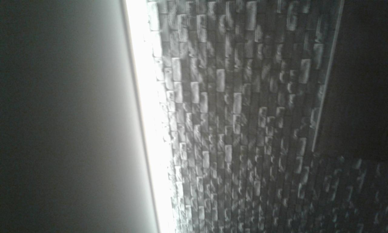 Ściana wykończona tapetą imitującą mur z cegieł w odcieniach szarości, oświetlona z boku światłem, z widocznym fragmentem gładkiej ściany po lewej stronie.