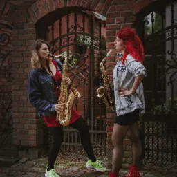 Dwie kobiety grają na saksofonach przed ceglanym łukiem z metalową bramą, jedna w czerwonej sukience i zielonych butach, druga w dżinsowej kurtce i czerwonych botkach.
