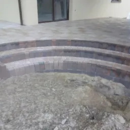 Budowa schodów zewnętrznych z kostki brukowej, widoczny etap układania pierwszych stopni w kształcie łuku, z przygotowanym podłożem. W tle elewacja budynku z oknami.