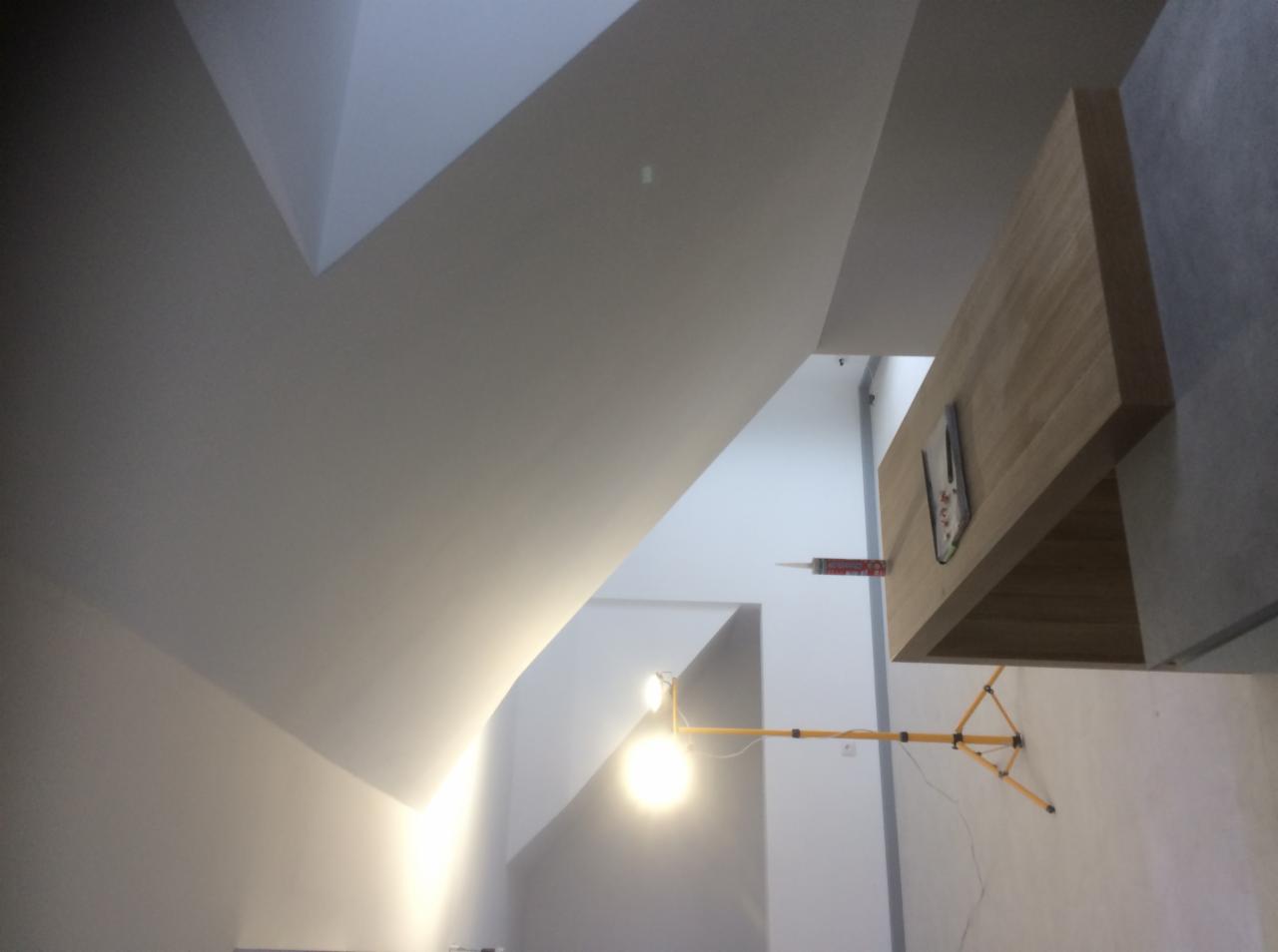 Wnętrze w trakcie prac wykończeniowych, widoczna lampa na statywie oświetlająca sufit z nietypowym załamaniem, obok ściany stoi blat z przyklejoną tubą silikonu.