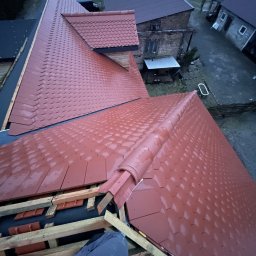SŁAWOMIR WOJTUSZKIEWICZ - Czerwony dach z dachówki karpiówki w trakcie układania, widok z góry na konstrukcję więźby i częściowo ułożone pokrycie. W tle zabudowania gospodarcze.