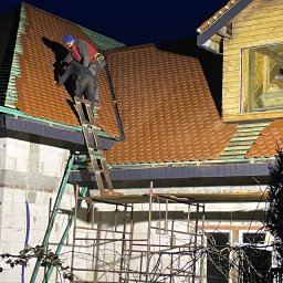 SŁAWOMIR WOJTUSZKIEWICZ - Ekipa dekarzy montuje brązową dachówkę na nowym dachu domu. Widoczne rusztowania i drabina. Praca wykonywana nocą, oświetlona sztucznym światłem.