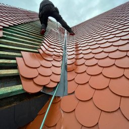 SŁAWOMIR WOJTUSZKIEWICZ - Układanie czerwonej dachówki karpiówki na drewnianej konstrukcji dachu. Widoczny dekarz w trakcie pracy, pochmurne niebo w tle. Dachówka błyszczy po deszczu.