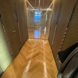 SŁAWOMIR WOJTUSZKIEWICZ - Korytarz z parkietem w jodełkę i ścianami o fakturze skóry. Widoczne oświetlenie LED i przeszklone drzwi z metalową ramą. Częściowo widoczny wieszak z ubraniami.