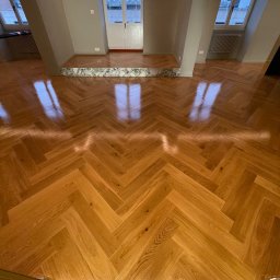 SŁAWOMIR WOJTUSZKIEWICZ - Parkiet ułożony w jodełkę w jasnym pomieszczeniu z oknami i marmurowym progiem. Widok na podłogę z perspektywy.