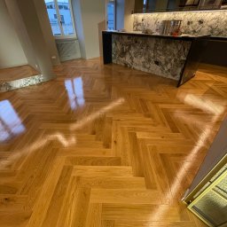 SŁAWOMIR WOJTUSZKIEWICZ - Parkiet ułożony w jodełkę w jasnym, nowoczesnym wnętrzu. Widoczny fragment marmurowego blatu i okno z widokiem na miasto. Podłoga po renowacji, z połyskiem.