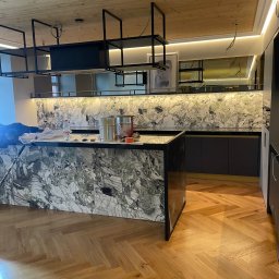 SŁAWOMIR WOJTUSZKIEWICZ - Nowoczesna kuchnia z marmurowym blatem i drewnianym sufitem. Widoczne metalowe półki i parkiet w jodełkę. Częściowo widoczne akcesoria malarskie na blacie.