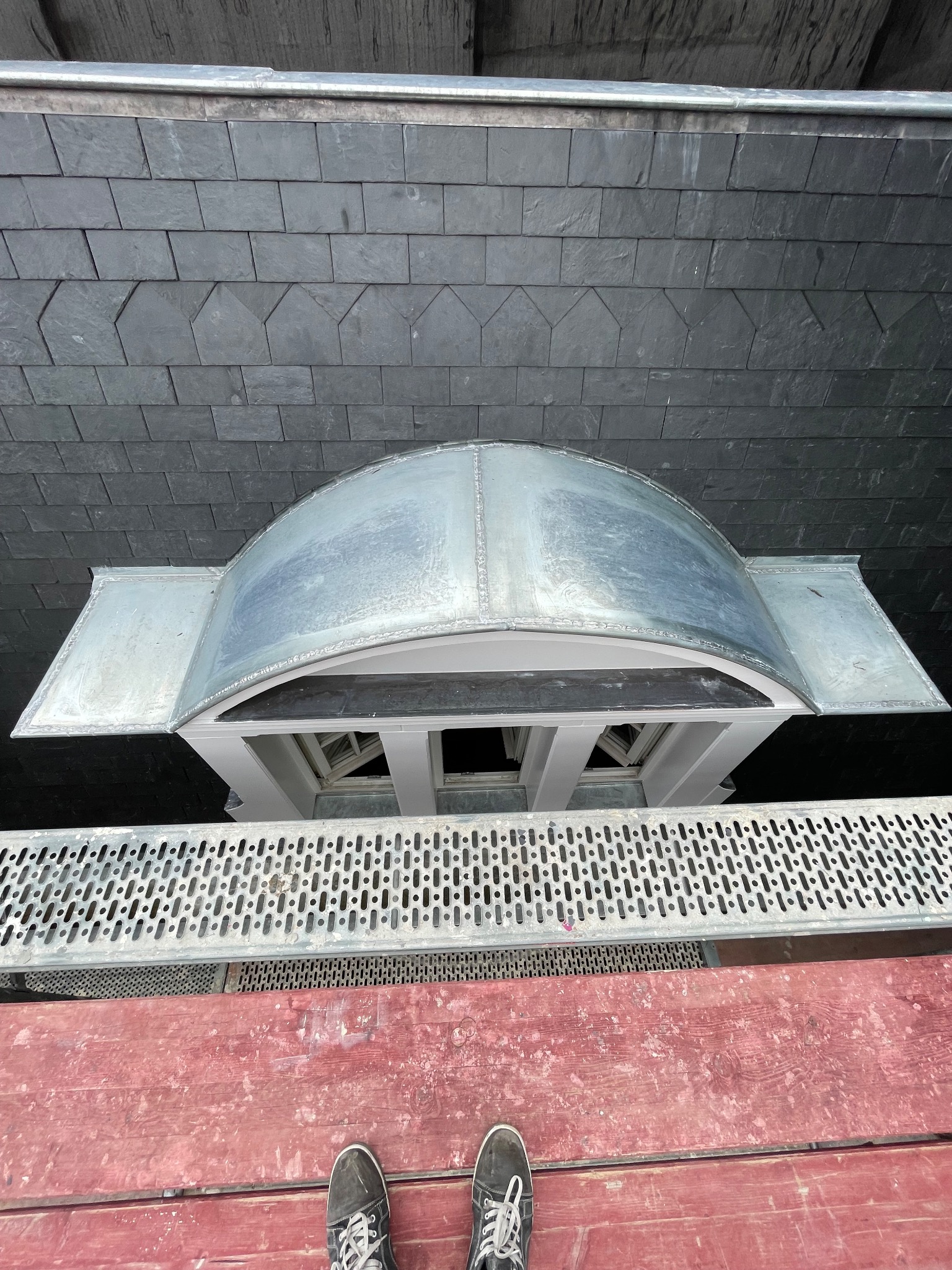 Widok z góry na fragment dachu pokrytego łupkiem, z metalowym obramowaniem okna dachowego i fragmentem rusztowania. U dołu widoczne stopy w obuwiu sportowym.