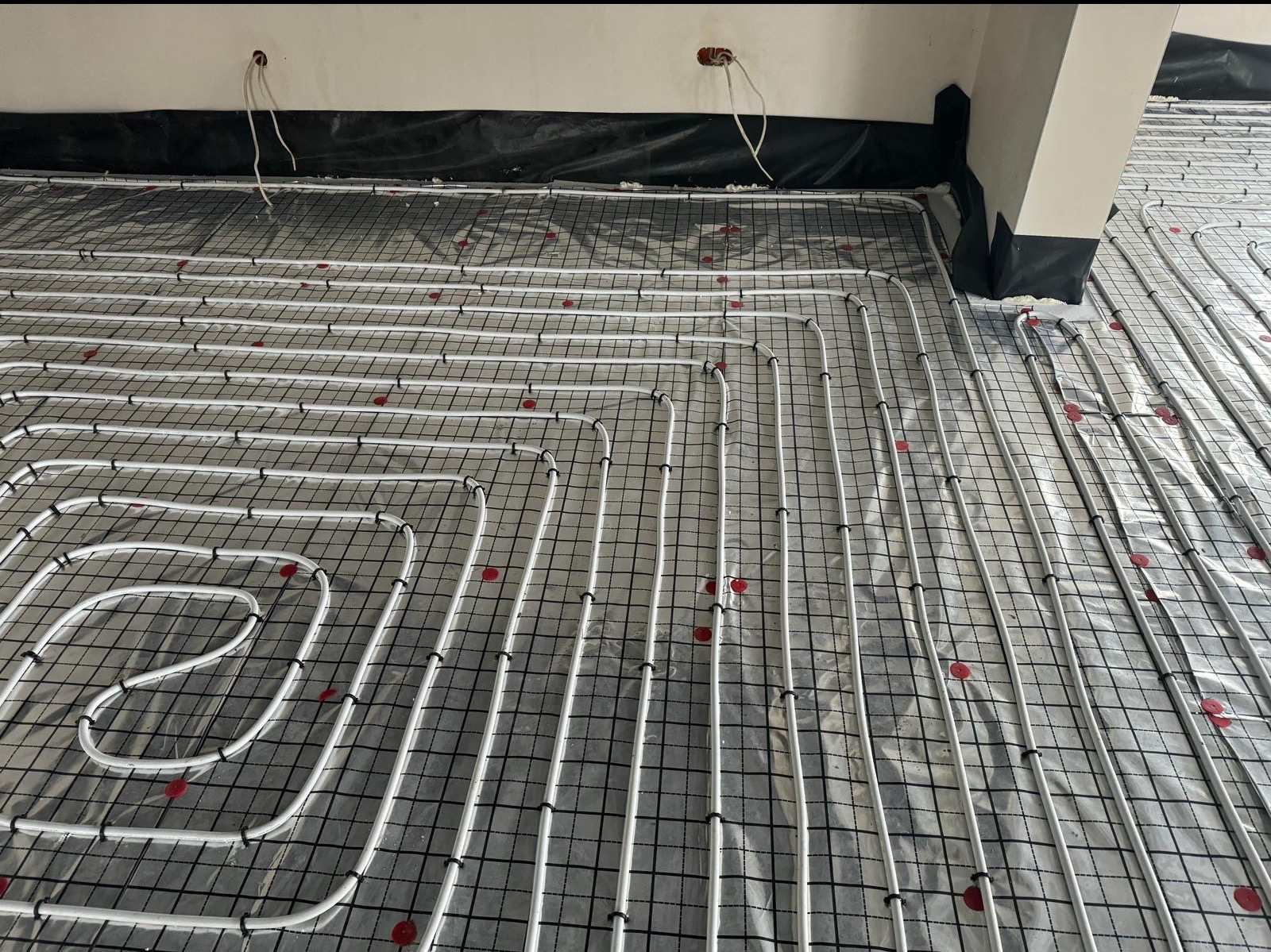 Białe rury ogrzewania podłogowego ułożone na czarnej siatce z czerwonymi znacznikami, widoczne kable elektryczne przy ścianie. Etap instalacji przed wylaniem posadzki.