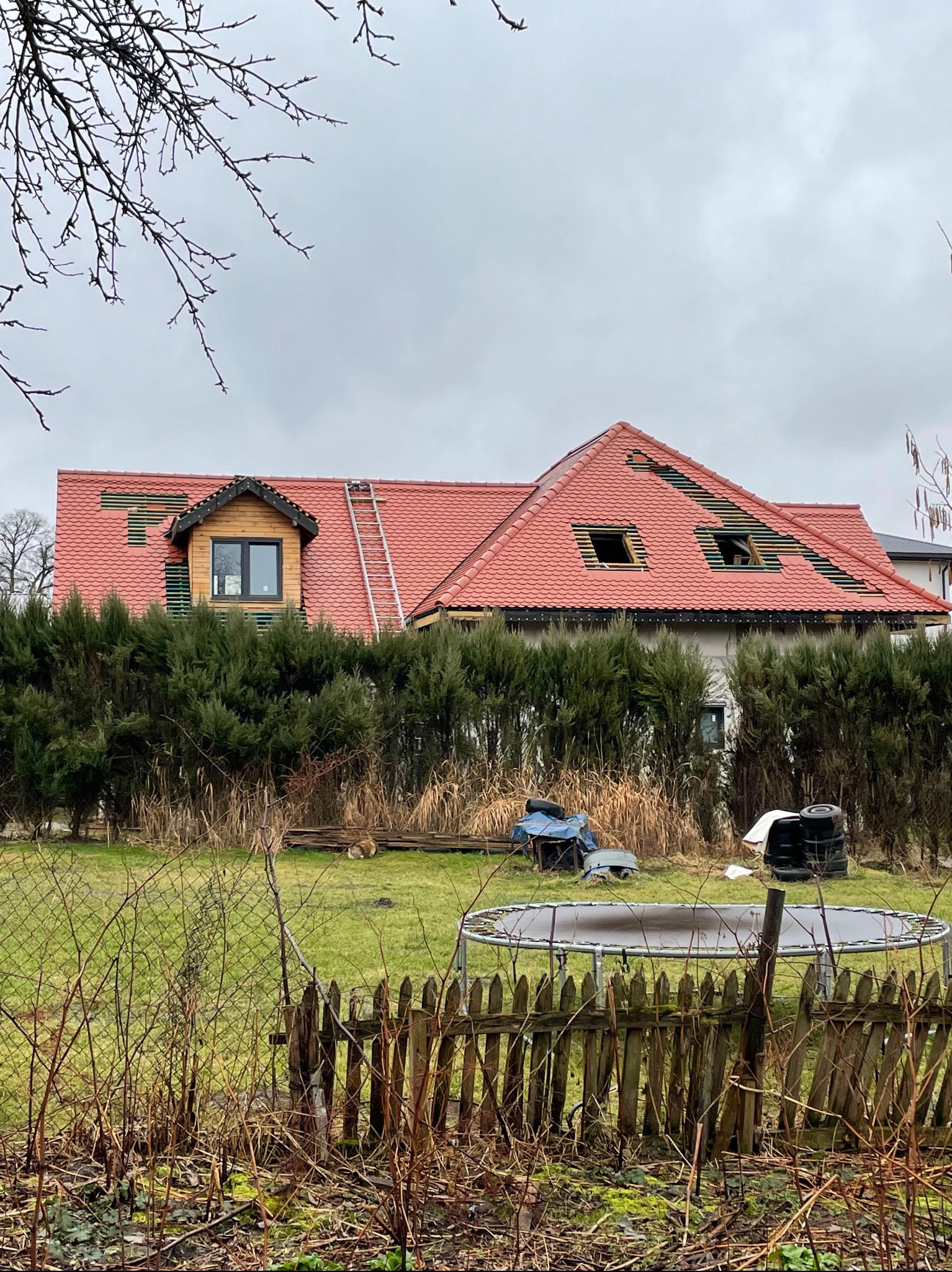 Czerwony dach z lukarną w trakcie wymiany, widoczny zza ogrodzenia w Urzędowie. Na dachu drabina, fragmenty łat. Tło stanowią krzewy i pochmurne niebo.