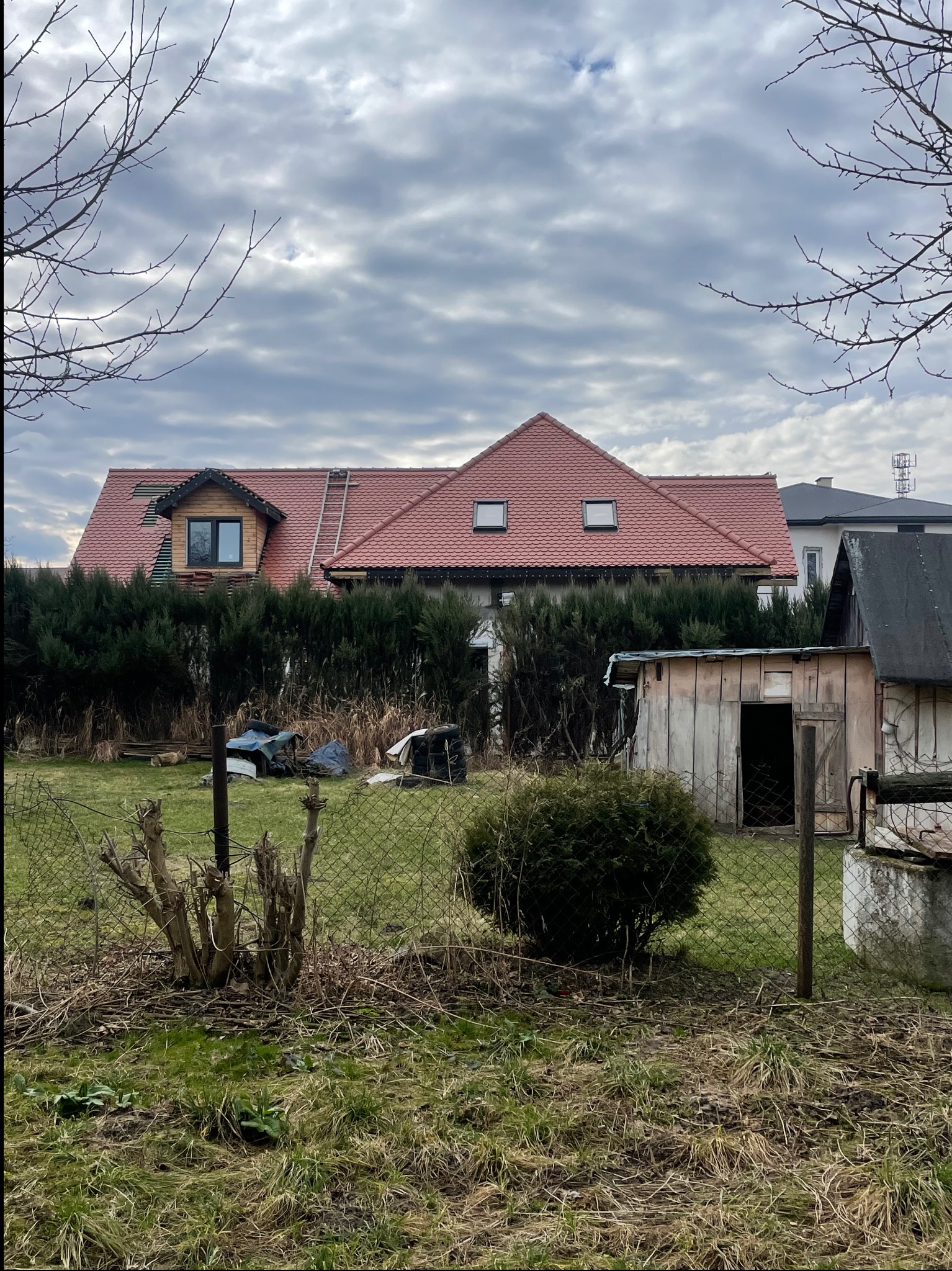 Dom z czerwonym dachem po wymianie, widoczny zza ogrodzenia i zieleni. Na dachu drabina, a w dachu okna. Pochmurne niebo.