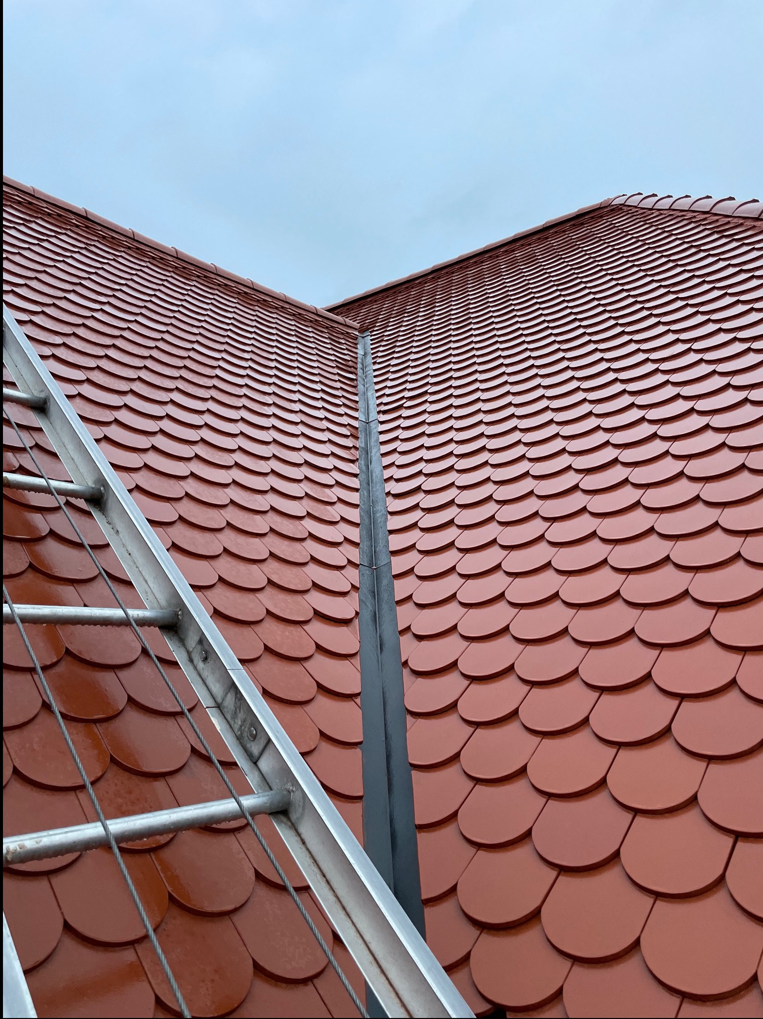 Czerwony dach z dachówki karpiówki, widoczny z dołu pod kątem, z fragmentem drabiny dekarskiej po lewej stronie. Dach jest nowy i błyszczący, a na niebie widać pochmurne niebo.