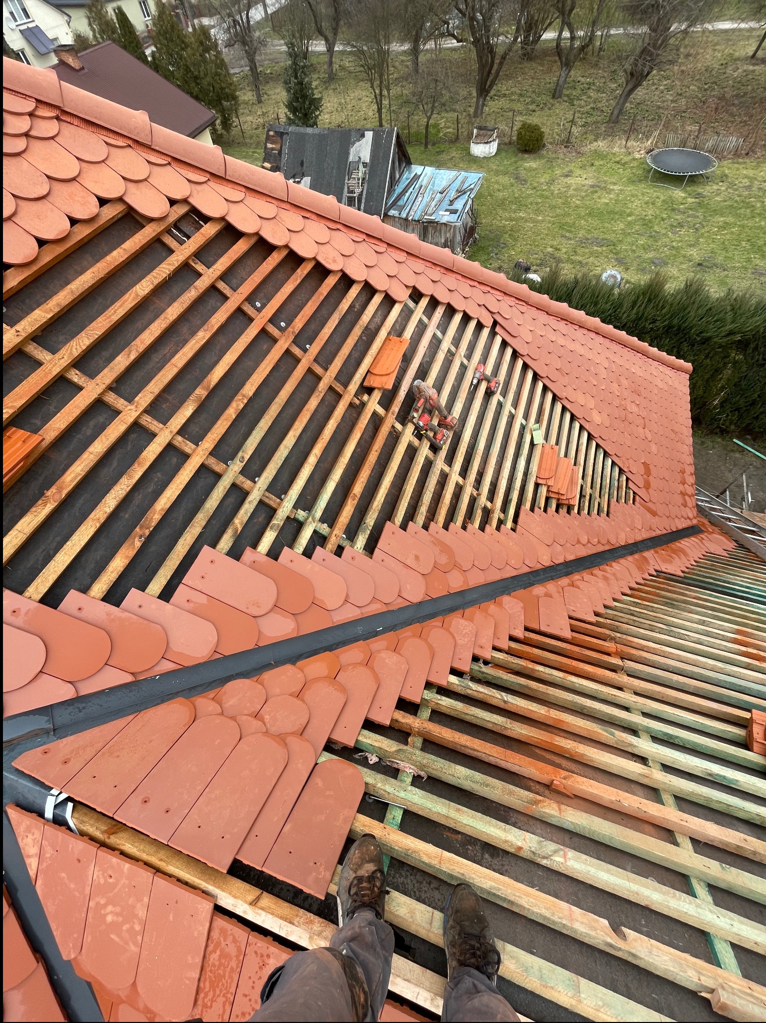 Częściowy widok dachu w trakcie krycia czerwoną dachówką karpiówką. Widoczne łaty, kontrłaty, membrana i narzędzia. Ujęcie z perspektywy osoby stojącej na dachu.