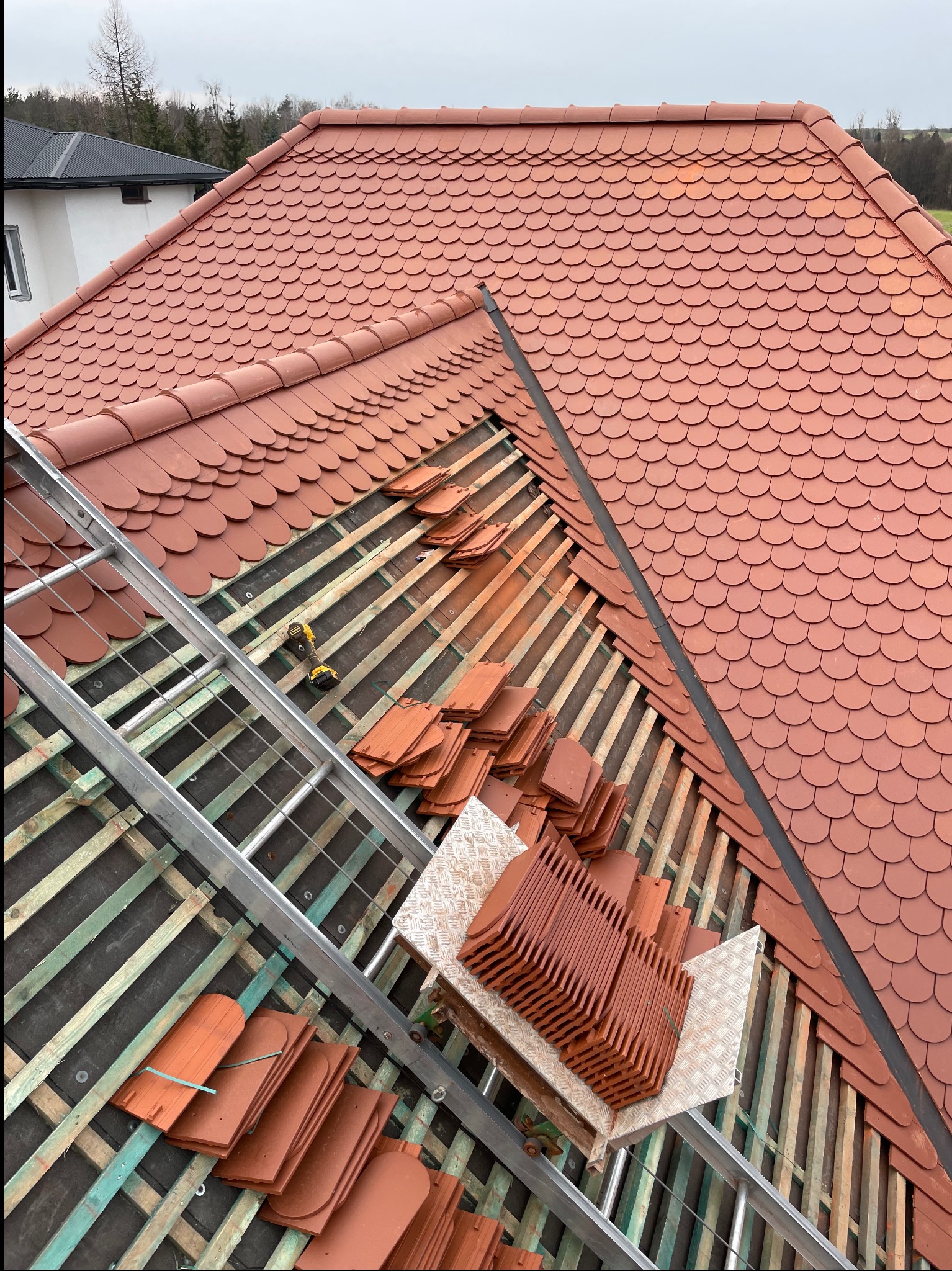 Częściowo pokryty dach z czerwonej dachówki karpiówki, widoczne łaty, drabina i narzędzia. Fragment dachu w trakcie układania, z bliska, z góry.