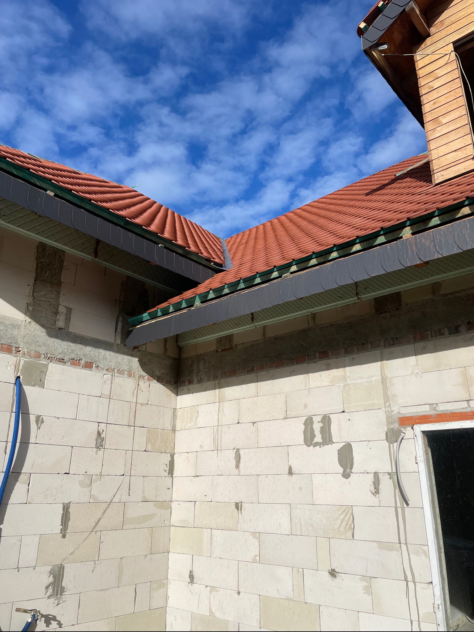 Wznoszenie ścian domu z bloczków, widoczny fragment dachu z czerwoną dachówką i szarymi elementami wykończeniowymi. Błękitne niebo w tle.