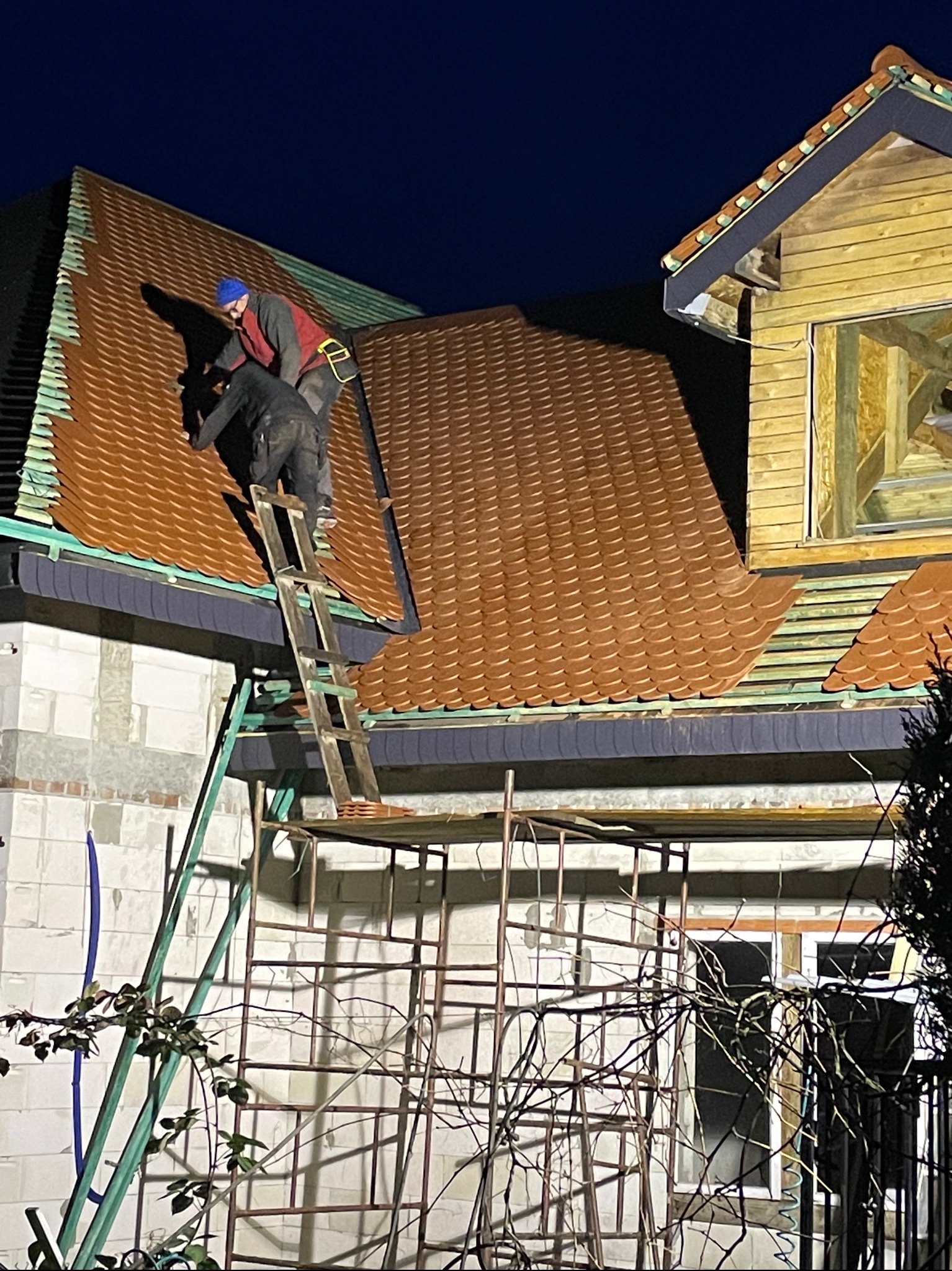 Ekipa dekarzy montuje brązową dachówkę na nowym dachu domu. Widoczne rusztowania i drabina. Praca wykonywana nocą, oświetlona sztucznym światłem.