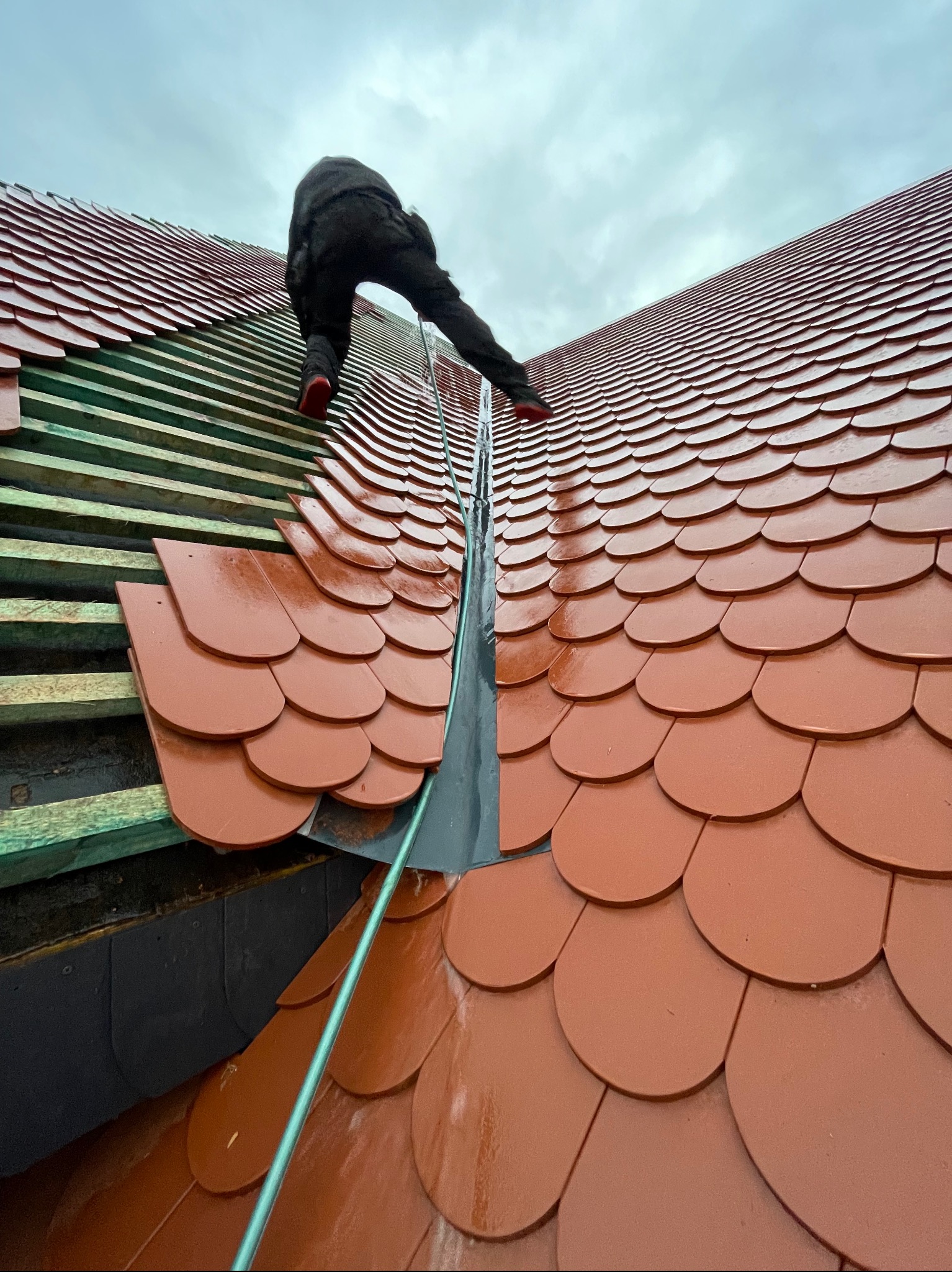 Układanie czerwonej dachówki karpiówki na drewnianej konstrukcji dachu. Widoczny dekarz w trakcie pracy, pochmurne niebo w tle. Dachówka błyszczy po deszczu.