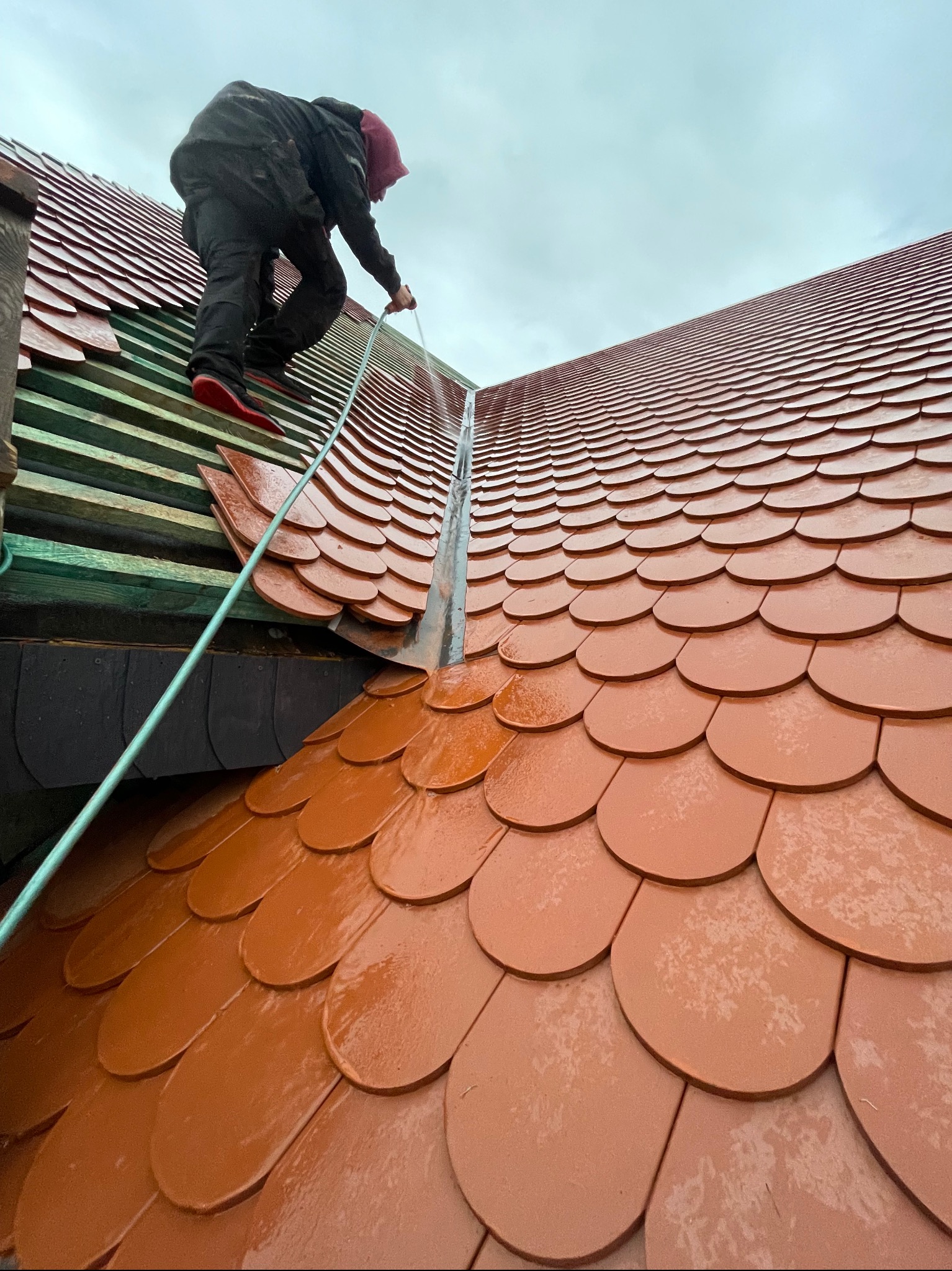 Pracownik na dachu z łuski ceramicznej, czyści rynnę. Widoczne drewniane elementy konstrukcji dachu i fragment nieba. Ujęcie z dołu, podkreślające stromość dachu.