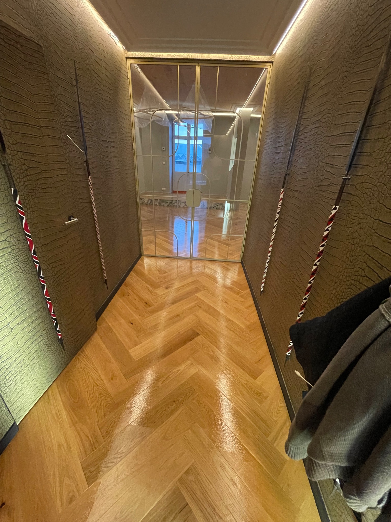 Korytarz z parkietem w jodełkę i ścianami o fakturze skóry. Widoczne oświetlenie LED i przeszklone drzwi z metalową ramą. Częściowo widoczny wieszak z ubraniami.