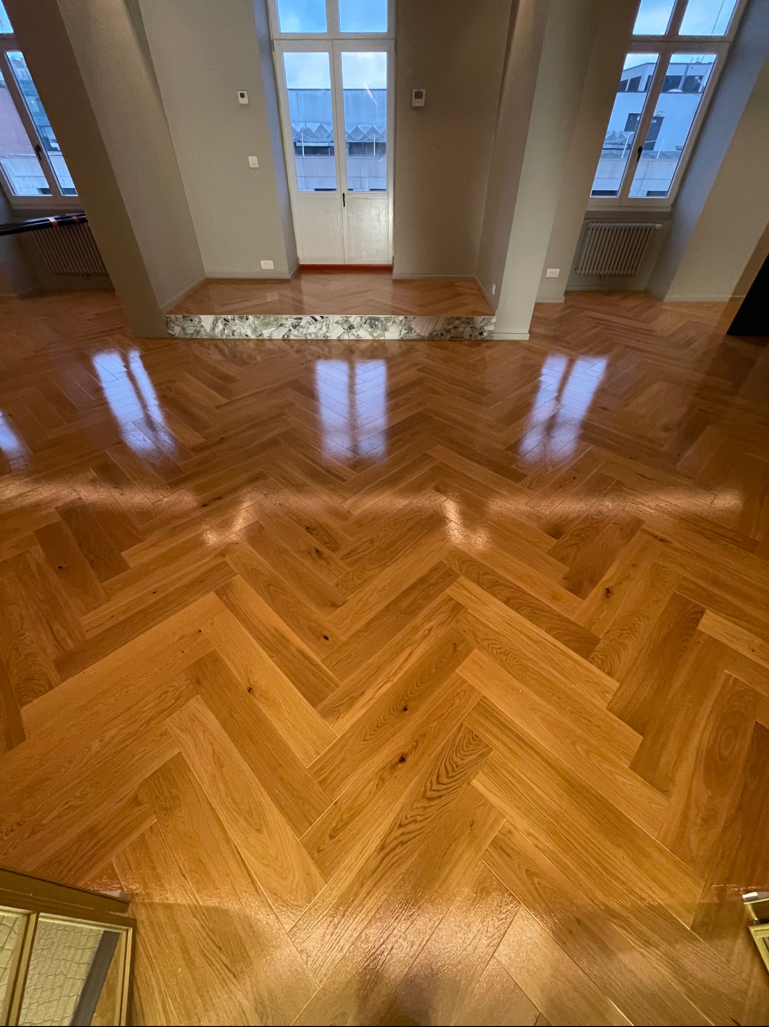 Parkiet ułożony w jodełkę w jasnym pomieszczeniu z oknami i marmurowym progiem. Widok na podłogę z perspektywy.