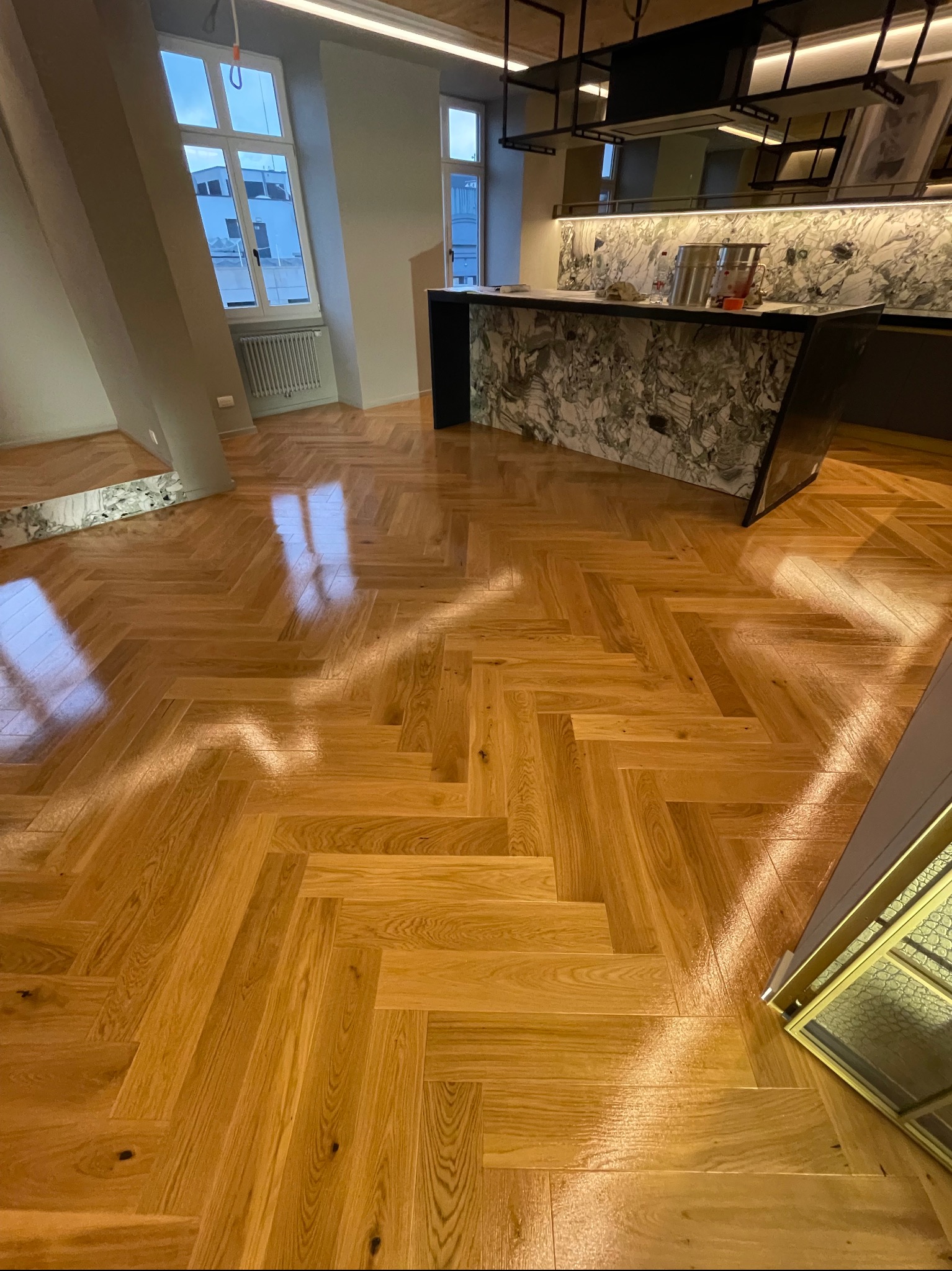 Parkiet ułożony w jodełkę w jasnym, nowoczesnym wnętrzu. Widoczny fragment marmurowego blatu i okno z widokiem na miasto. Podłoga po renowacji, z połyskiem.