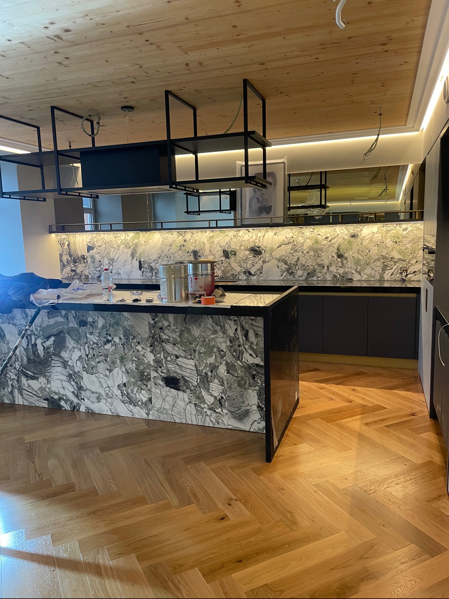 Nowoczesna kuchnia z marmurowym blatem i drewnianym sufitem. Widoczne metalowe półki i parkiet w jodełkę. Częściowo widoczne akcesoria malarskie na blacie.