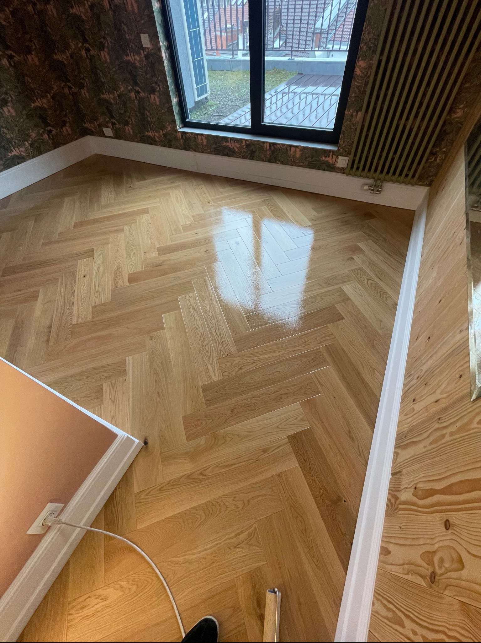 Parkiet ułożony w jodełkę francuską, świeżo po lakierowaniu, z widocznym odbiciem światła z okna. Dekoracyjna tapeta na ścianie i listwy przypodłogowe.