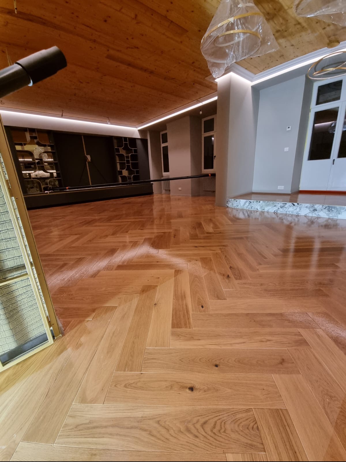 Parkiet ułożony w jodełkę w przestronnym wnętrzu z drewnianym sufitem i szarymi ścianami. Widoczne elementy oświetlenia i regały w tle. Podłoga po renowacji, gotowa do użytku.