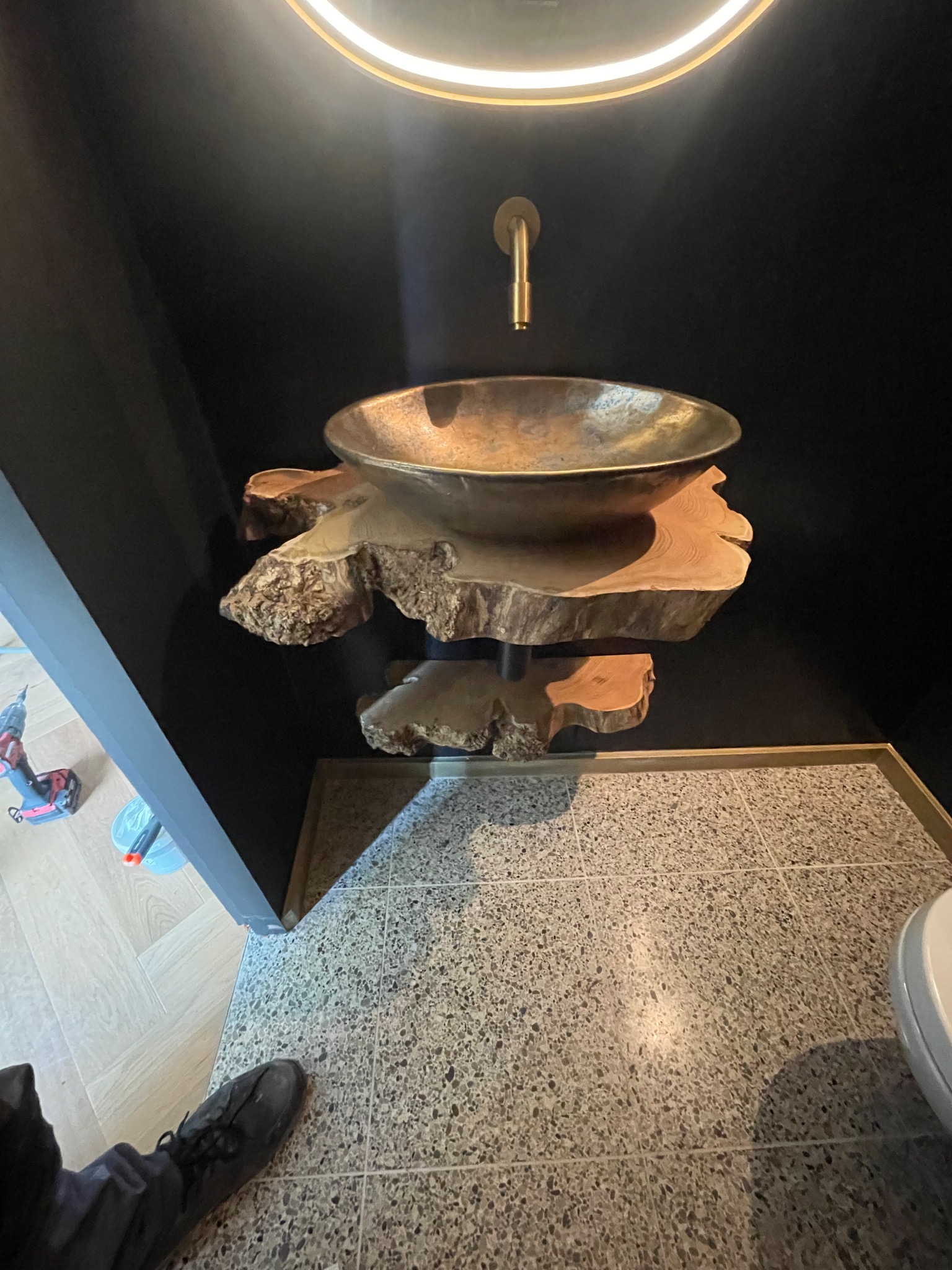 Nowoczesna łazienka z umywalką w stylu rustykalnym na drewnianym blacie, złota armatura i czarne ściany. Widoczny fragment podłogi z terazzo.