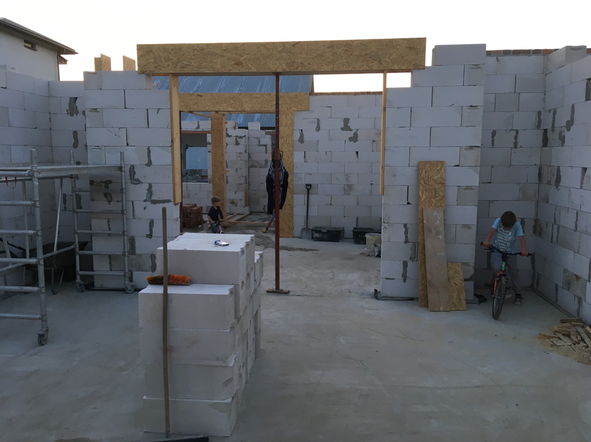 Wnętrze surowego domu z betonu komórkowego w trakcie budowy. Widoczne ściany, otwory okienne i drzwiowe, oraz materiały budowlane. Chłopiec na rowerze w tle.