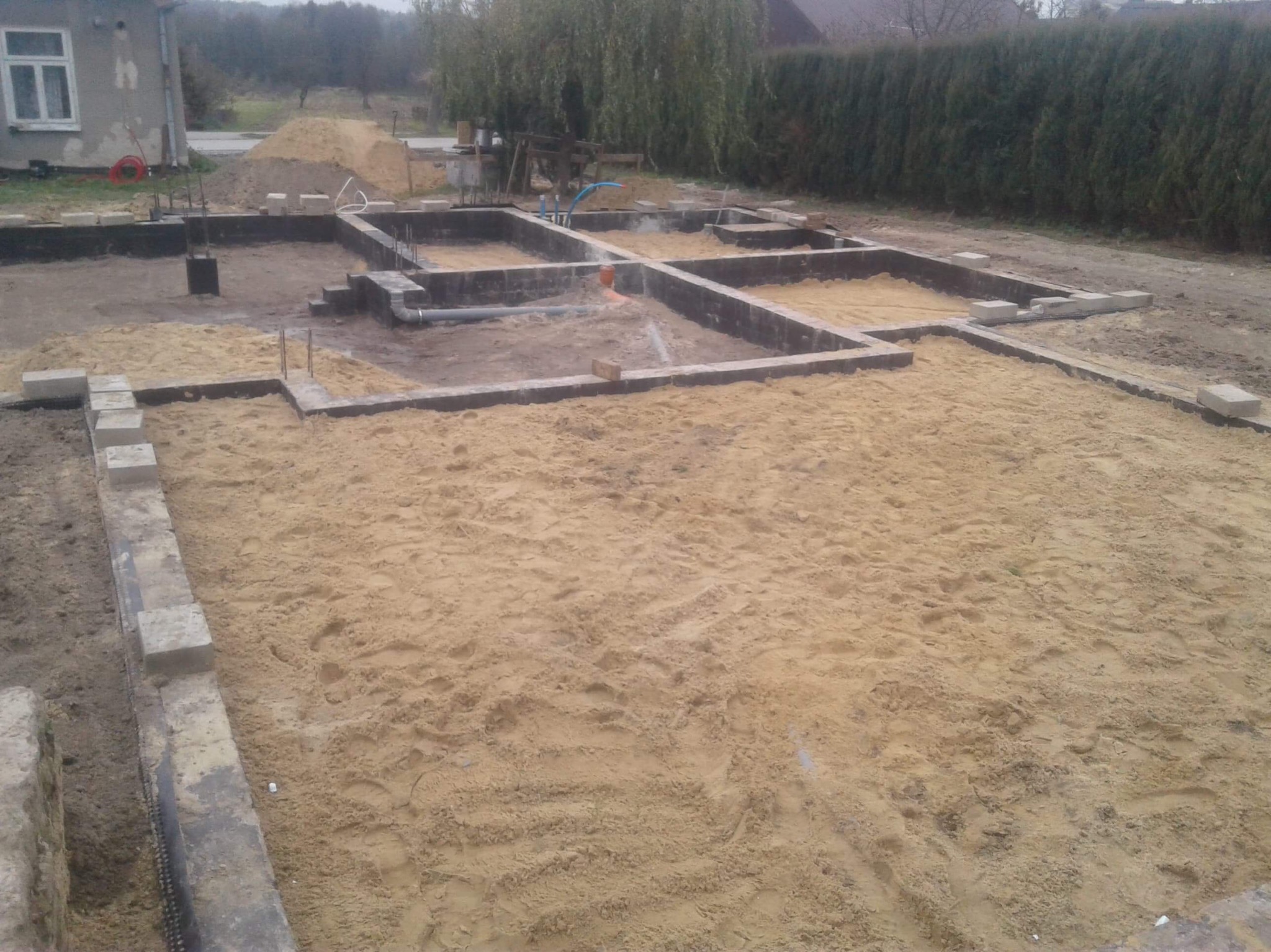 Fundamenty domu w trakcie budowy: zbrojenie, szalunki i piasek. Widoczne elementy instalacji. Ujęcie z góry, na pierwszym planie piasek.